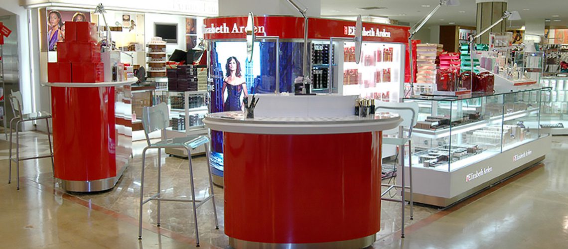 A brightly lit cosmetics store with a prominent red display and several makeup products on counters. A large poster of a woman is visible in the background. Shelves are stocked with beauty products.